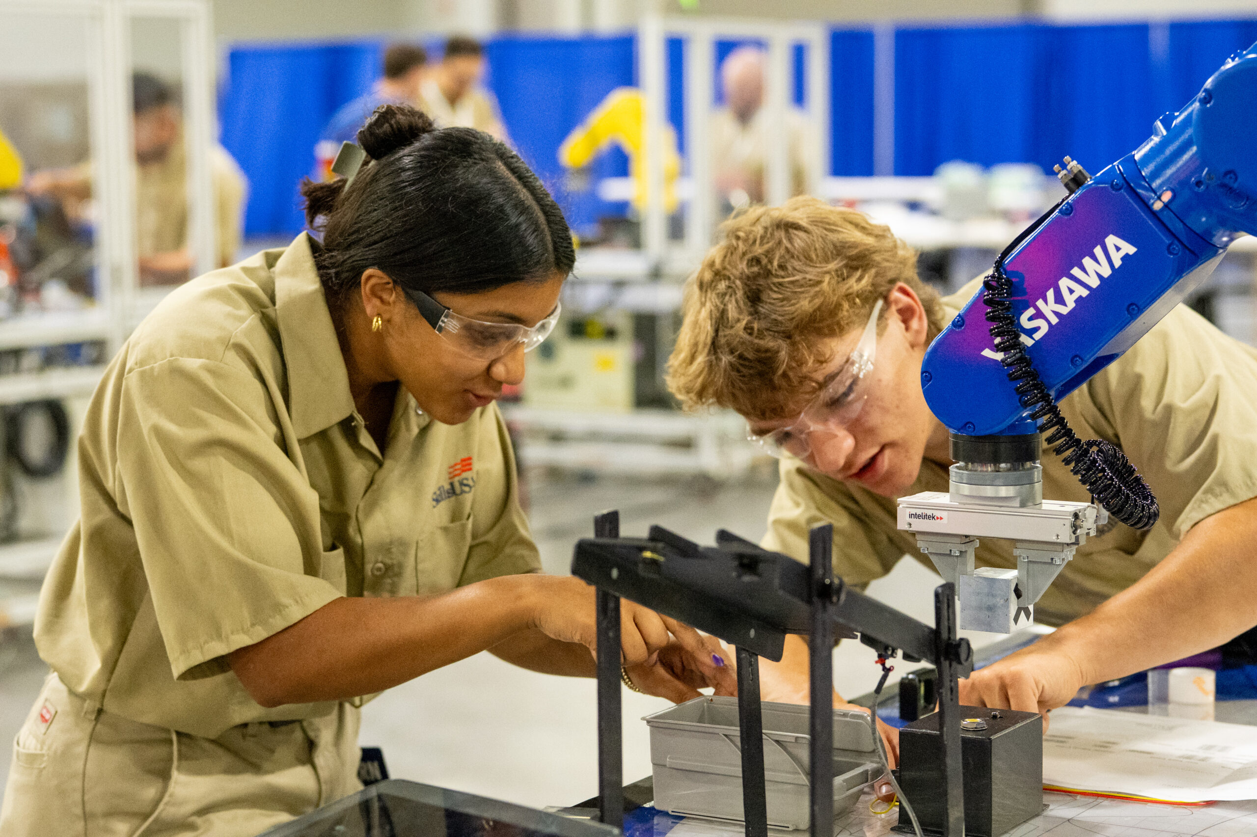 Two individuals in safety goggles and work uniforms collaborate with a robotic arm at a manufacturing station.