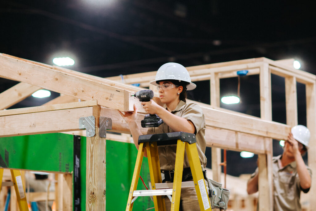 A construction worker uses a power drill to secure wooden beams while standing on a ladder inside a building under construction.