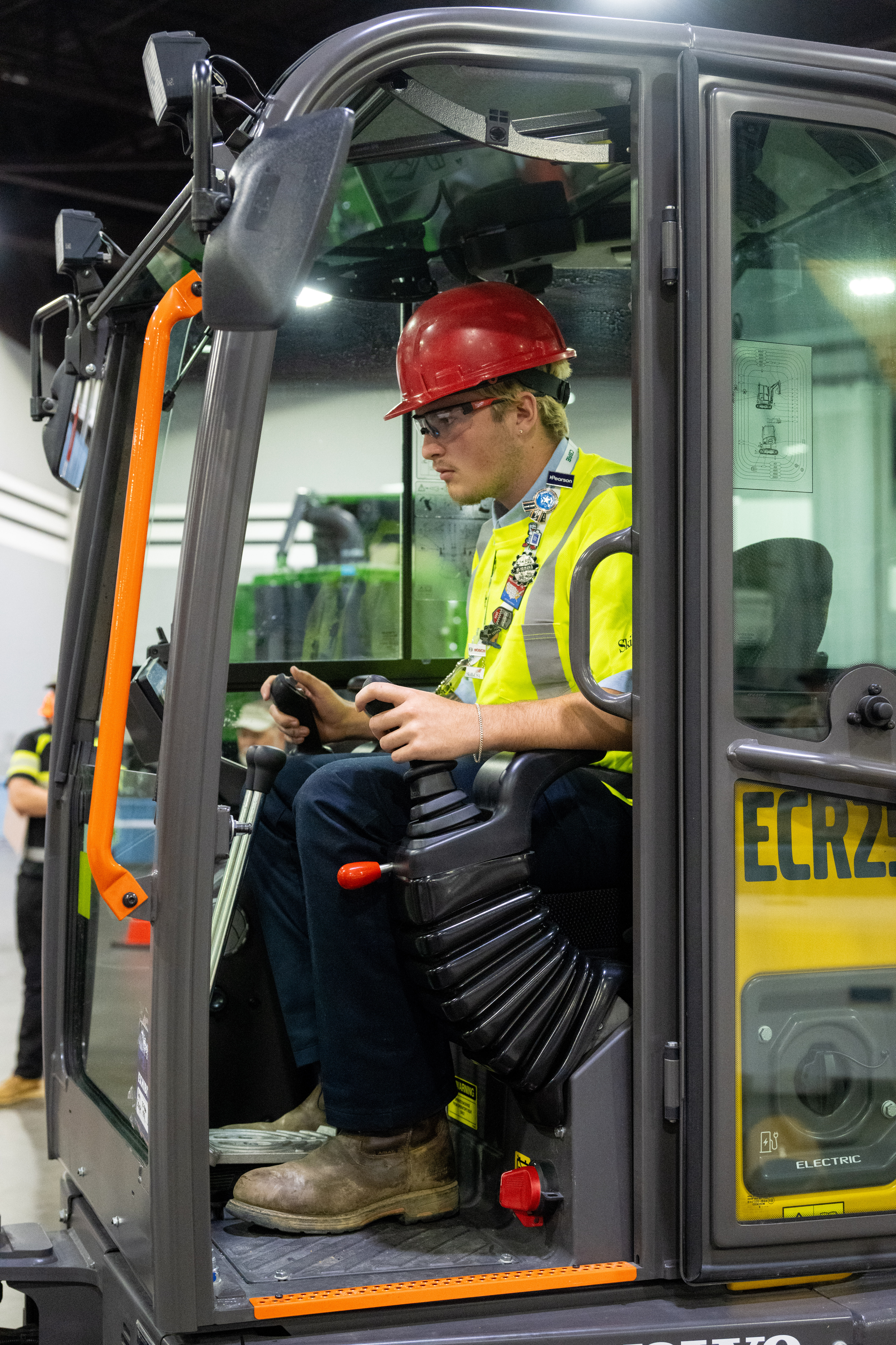 A person in a red hard hat and high-visibility vest operates an electric construction vehicle inside a facility.