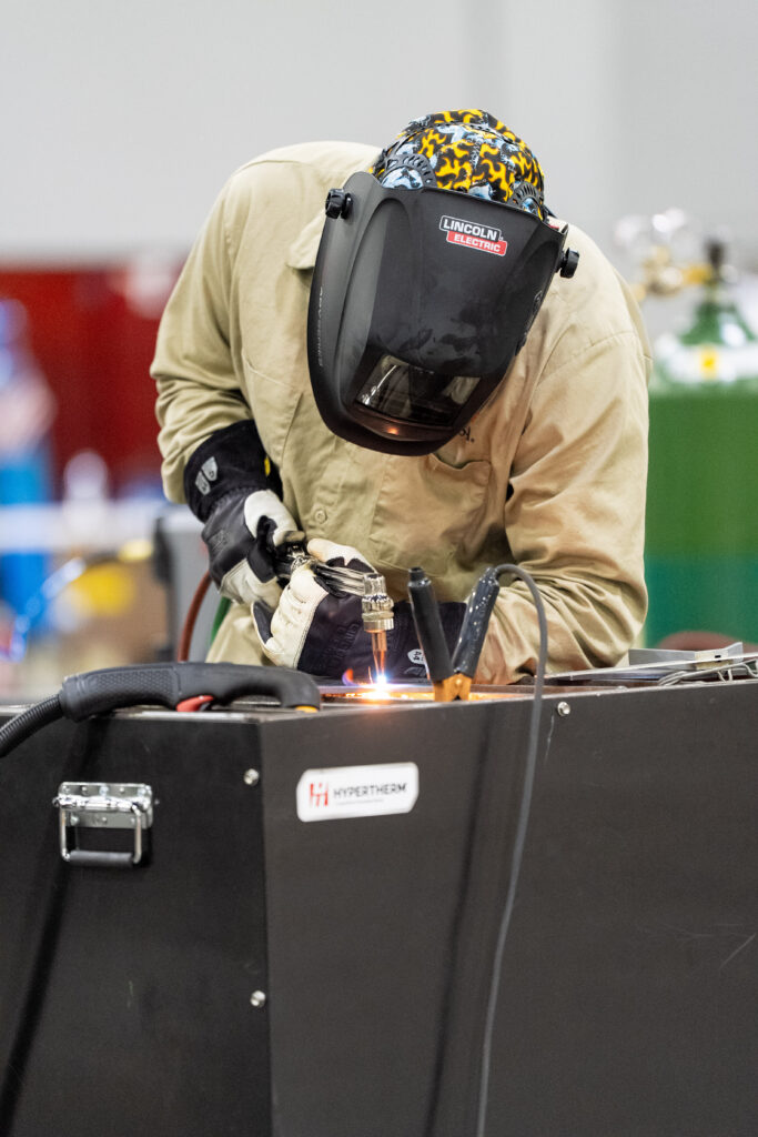 A welder in protective gear operates a welding torch on a metal surface, with sparks flying as they work.