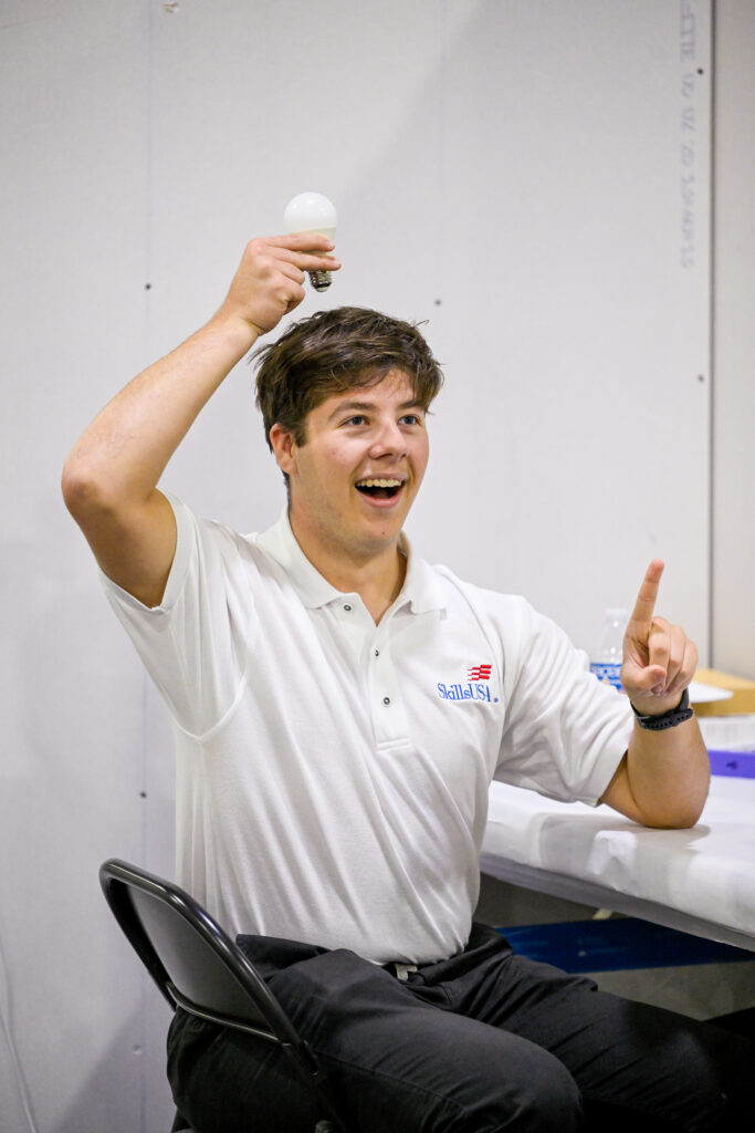 A person in a white polo shirt with a "SkillsUSA" logo sits in a chair, holding a light bulb above their head while pointing upward with their other hand.