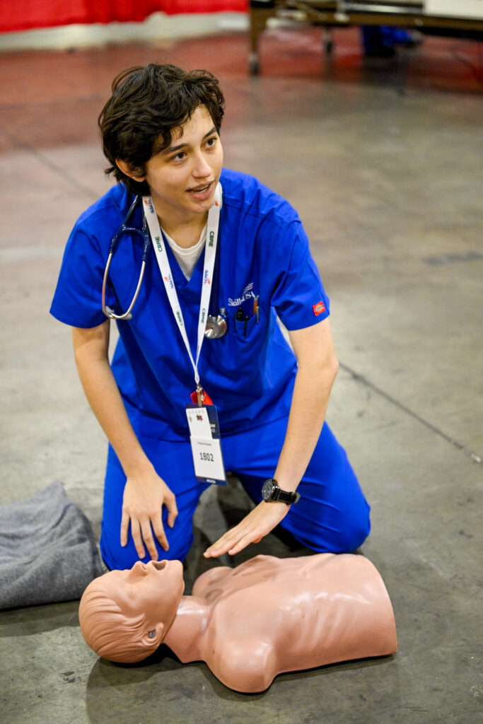 A person in blue scrubs kneels beside a CPR training mannequin, preparing to demonstrate rescue techniques.