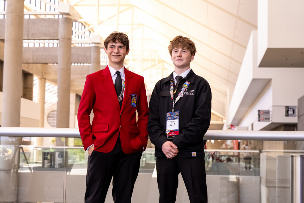 Two young individuals stand together in a modern building, one wearing a red blazer and the other in a black jacket with a lanyard and badge.