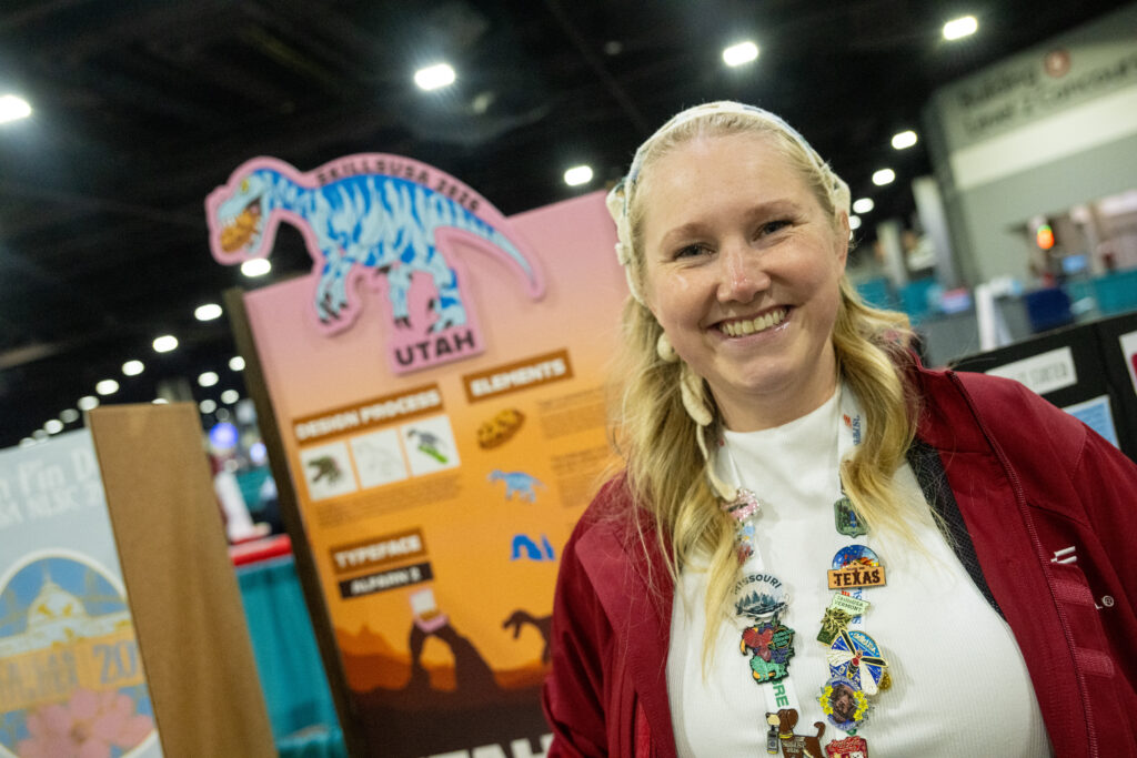 A smiling person with blonde hair and a headband stands at a convention booth featuring a colorful dinosaur display, adorned with numerous state-themed pins.