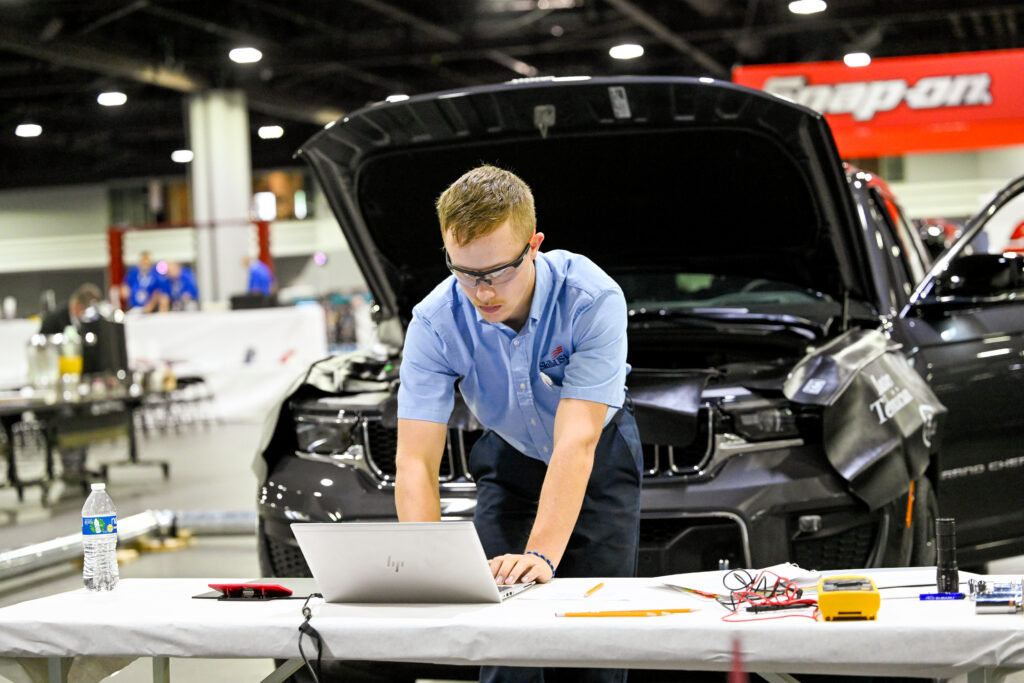 A technician leans over a laptop while working on a damaged black vehicle at an auto repair event.