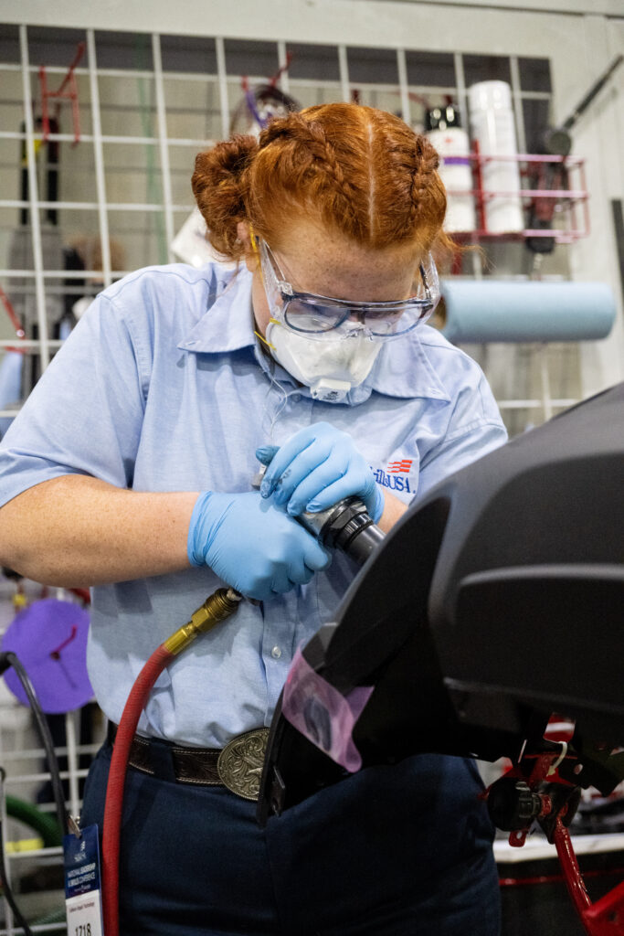 A person with red hair, wearing protective gear, uses a tool to work on a mechanical component in a workshop setting.